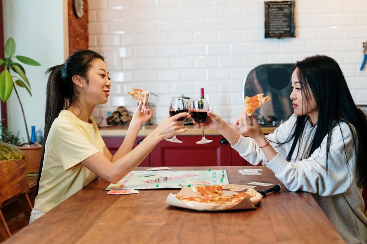 Two young female presenting people eat pizza over a board game